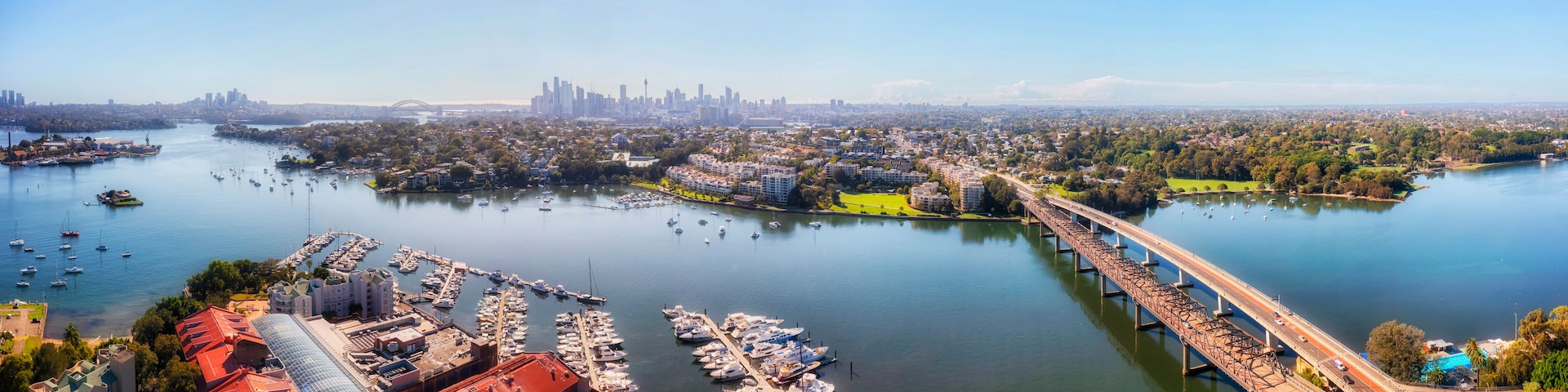 Un paisaje urbano costero con un puerto deportivo, un puente y un horizonte de la ciudad a lo lejos.
