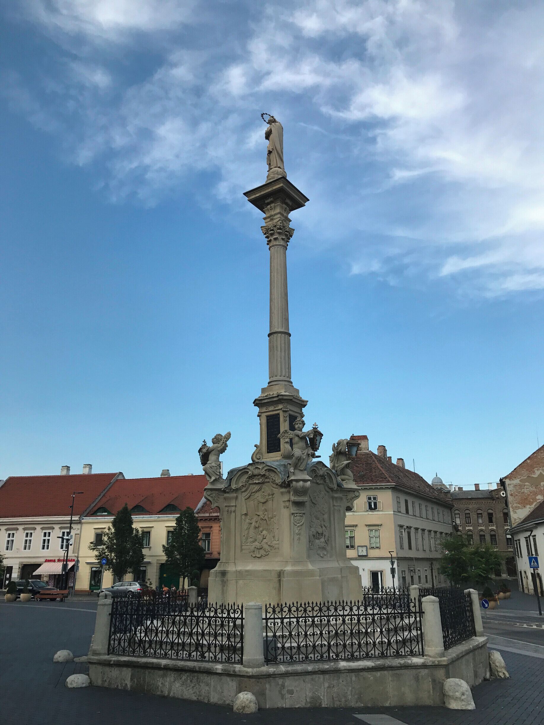 Um monumento alto com uma estátua no topo, cercado por uma grade de ferro forjado, com edifícios históricos ao fundo.