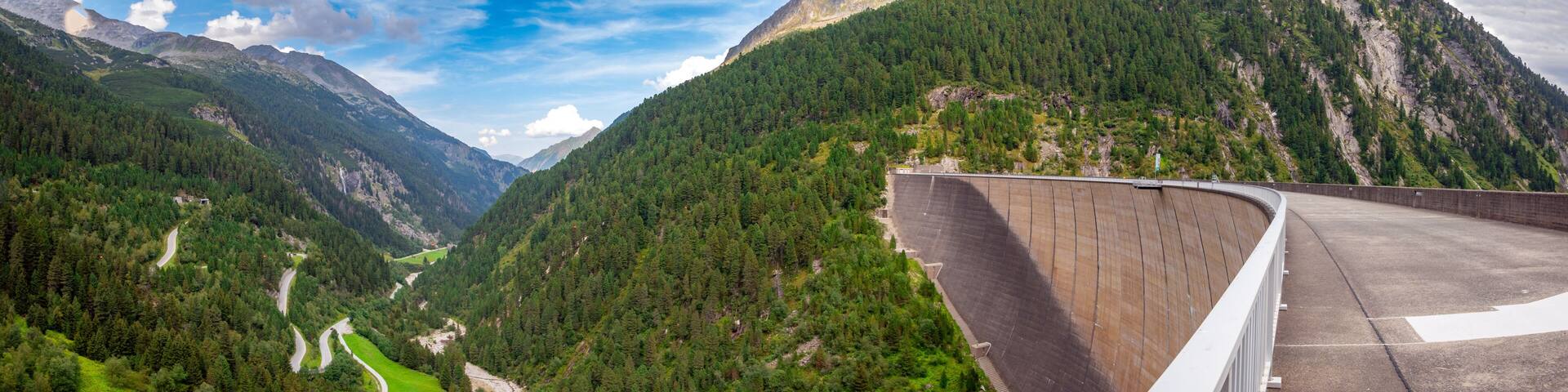 A dam, a winding road, and a valley with dense forests and mountains.