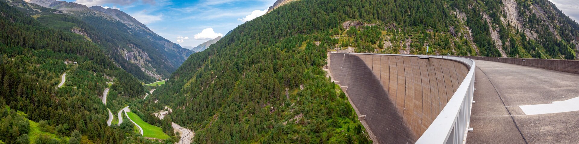 A dam, a winding road, and a valley with dense forests and mountains.