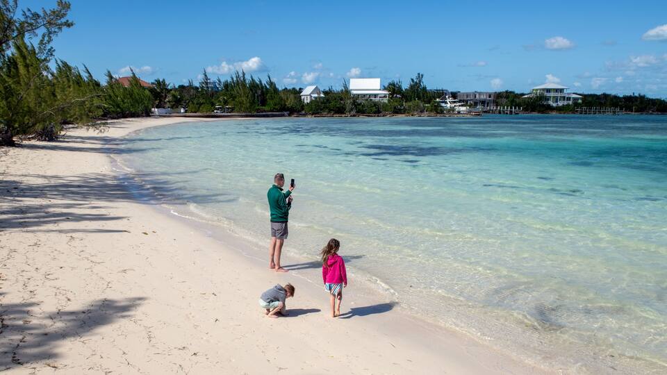 A sandy beach with clear turquoise water, a few people, and houses in the background.