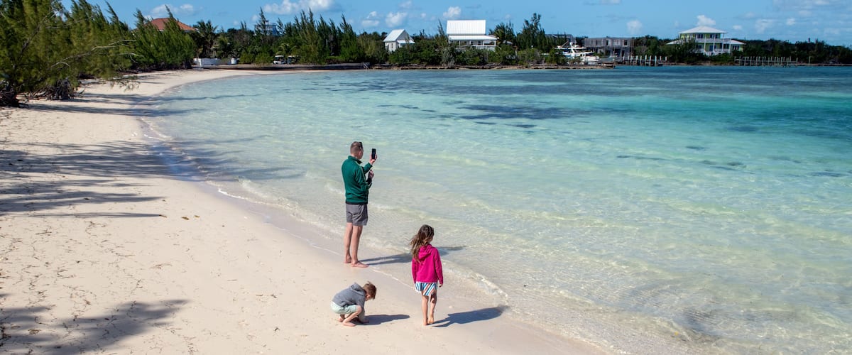 A sandy beach with clear turquoise water, a few people, and houses in the background.