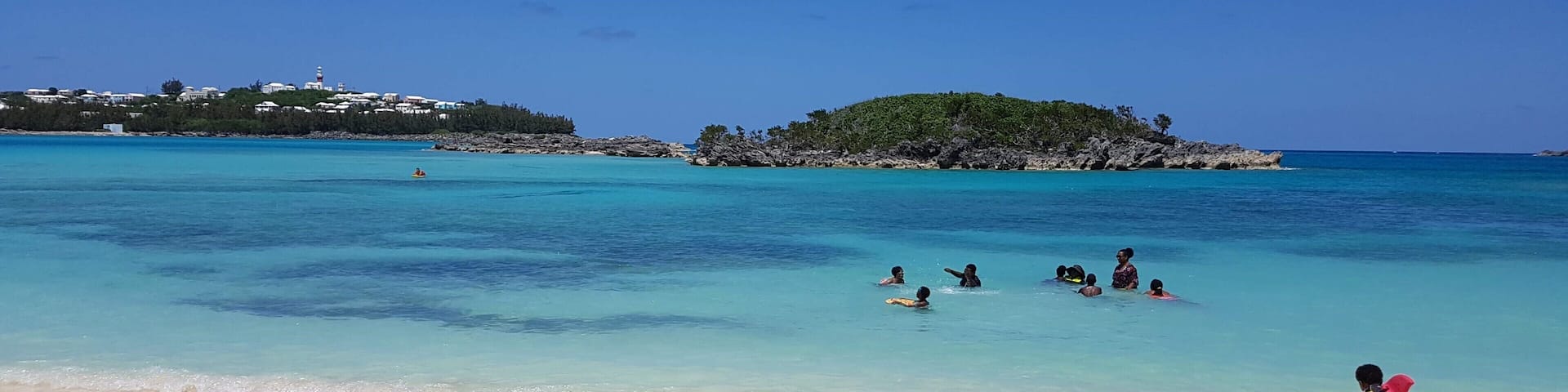 A beach with clear turquoise water, a sandy shore, and a small island with rocky terrain in the background.