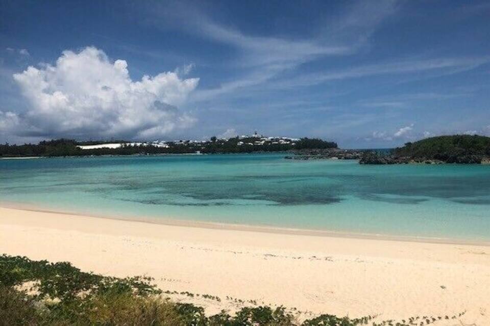 En strand med krystalklart turkis vand, en sandstrand og en fjerntliggende ø med bygninger.