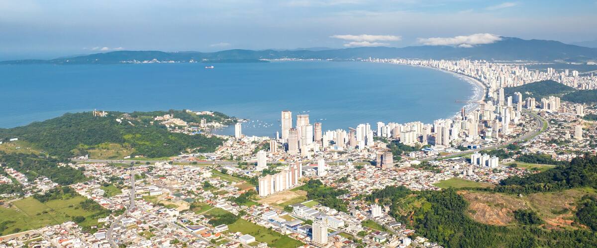 A coastal cityscape with dense urban development, a curved coastline, and surrounding green hills.
