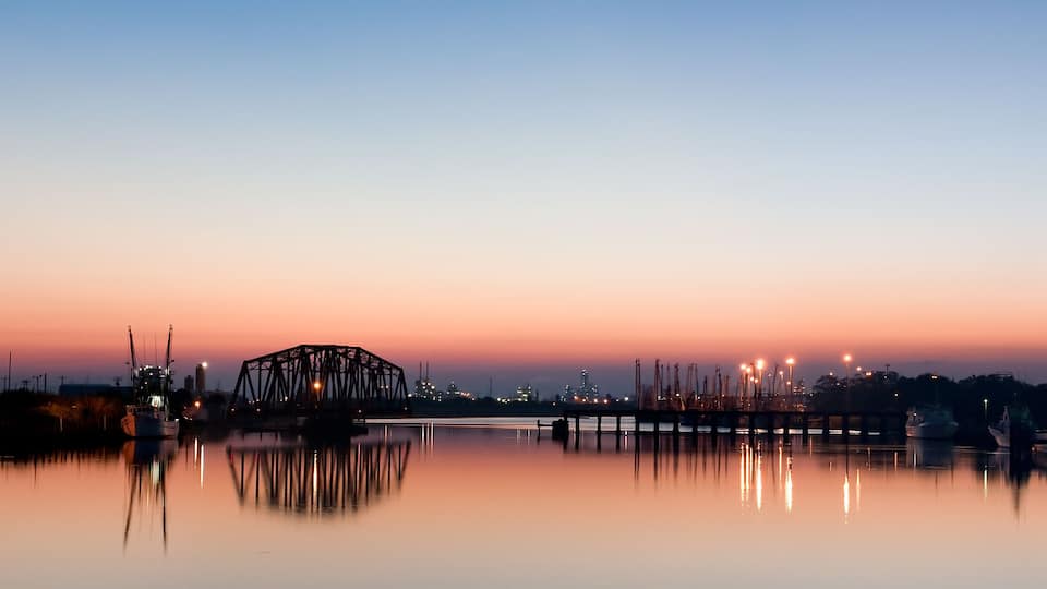 A calm harbor at dusk with a bridge, boats, and industrial structures in the background.