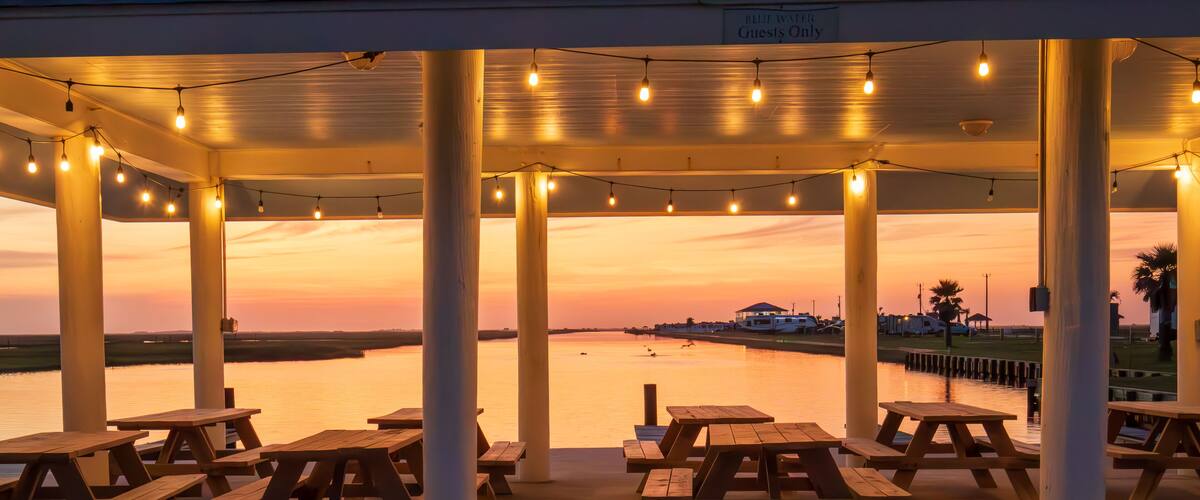 Picnic tables are neatly arranged under a covered pavilion adorned with hanging lights, providing a prime spot to enjoy the serene sunset over the calm waters in Freeport Texas.