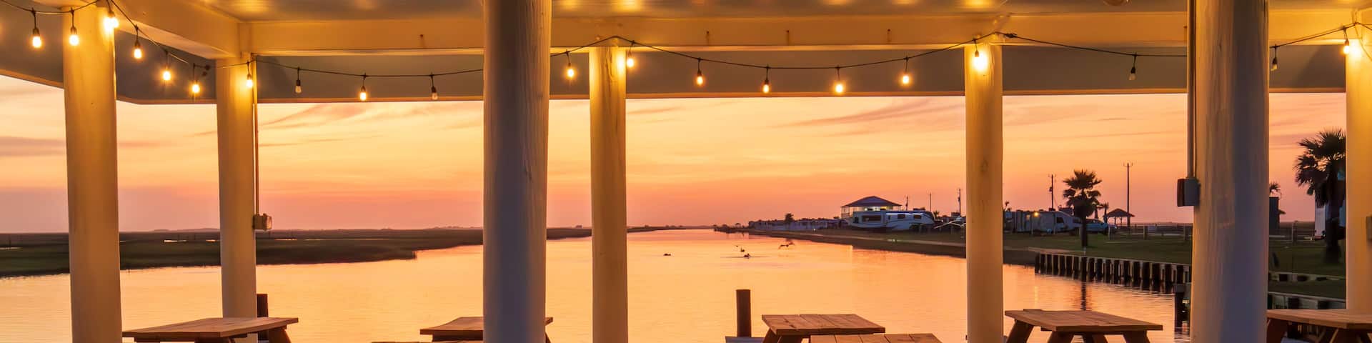 Picnic tables are neatly arranged under a covered pavilion adorned with hanging lights, providing a prime spot to enjoy the serene sunset over the calm waters in Freeport Texas.