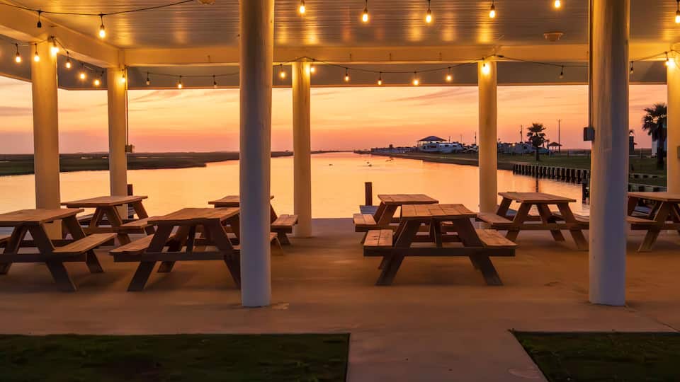 Picnic tables are neatly arranged under a covered pavilion adorned with hanging lights, providing a prime spot to enjoy the serene sunset over the calm waters in Freeport Texas.