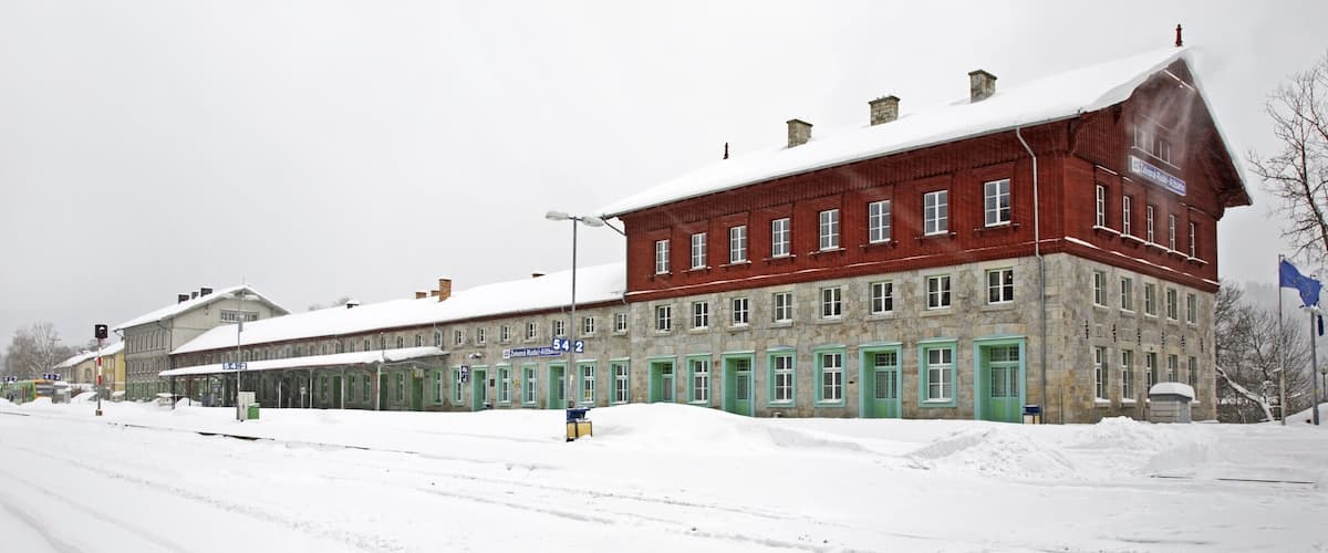 A red brick building with green doors and windows, surrounded by snow-covered ground and a cloudy sky.
