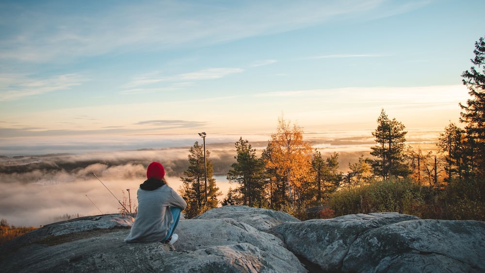 Hiker sits atop Vuokatinvaara in Sotkamo in the Kainuu region, Finland at sunrise and above the clouds. A brunette woman with a red knitted hat on top of the mountain