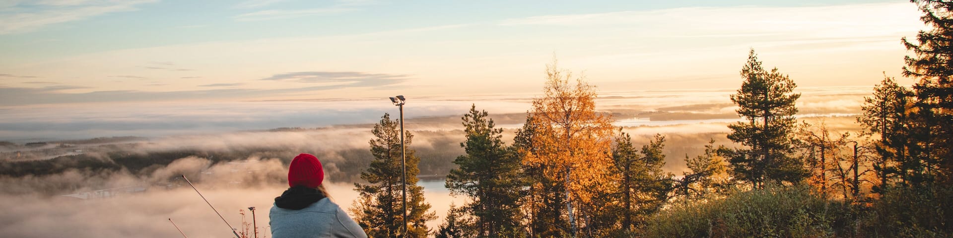 Hiker sits atop Vuokatinvaara in Sotkamo in the Kainuu region, Finland at sunrise and above the clouds. A brunette woman with a red knitted hat on top of the mountain