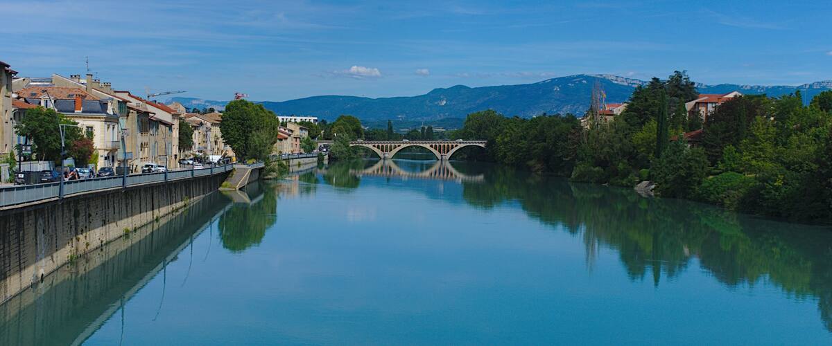 A calm river with a stone bridge, buildings along the riverbank, and mountains in the background.
