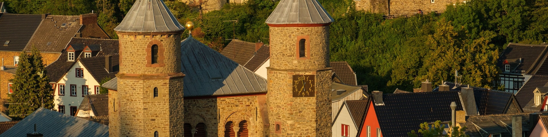 Uma igreja histórica com duas torres, um edifício de pedra com um relógio e um café-restaurante ao fundo.