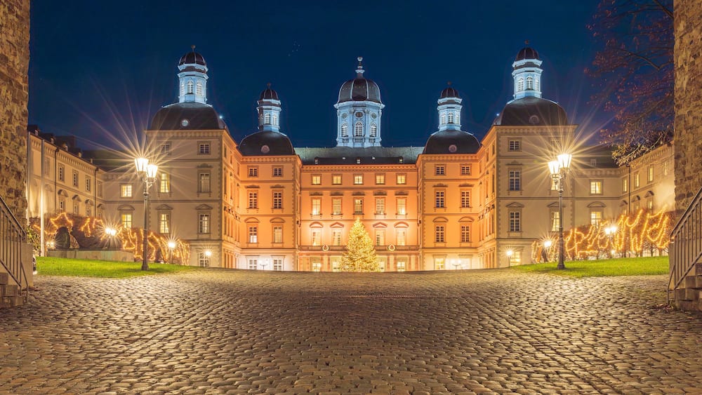 Un bâtiment historique doté de plusieurs dômes et de tours, une cour pavée et des arbres illuminés.