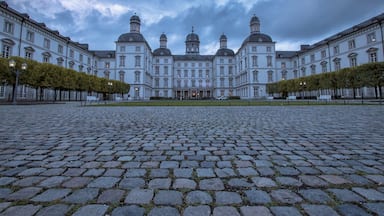 Een groot, historisch gebouw met meerdere koepels en symmetrische architectuur, omgeven door een geplaveid erf en omzoomd door bomen.