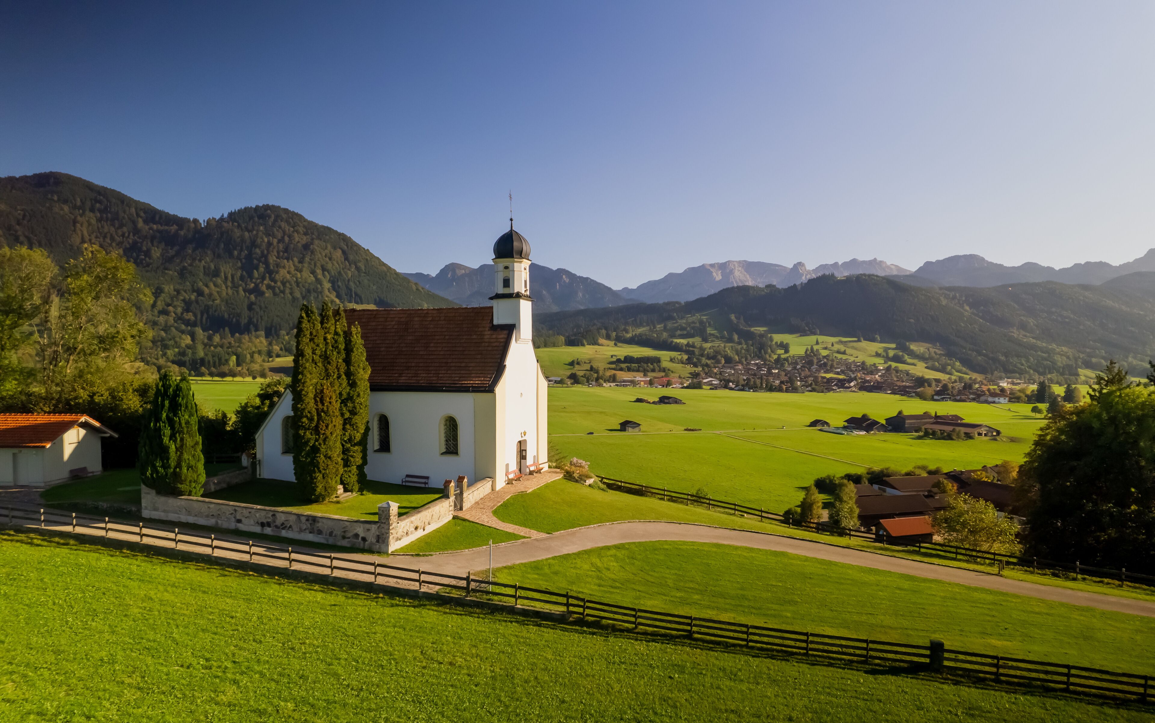 A white church with a bell tower, surrounded by green fields and mountains in the background.