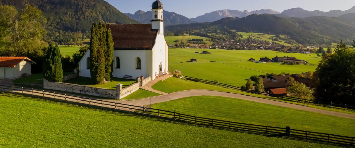 A white church with a bell tower, surrounded by green fields and mountains in the background.