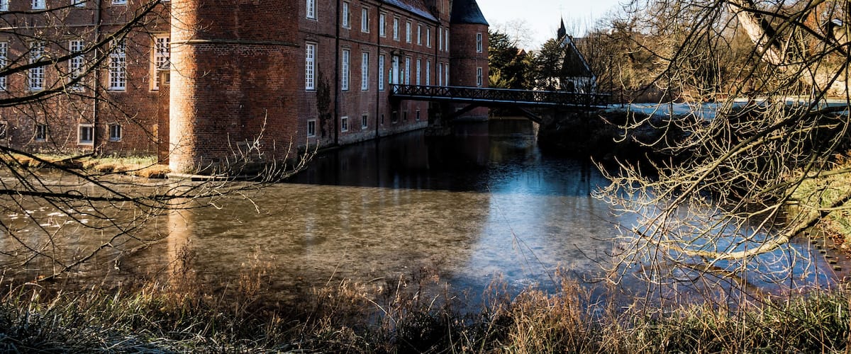 A historic brick building with a tower, a bridge over a river, and leafless trees.