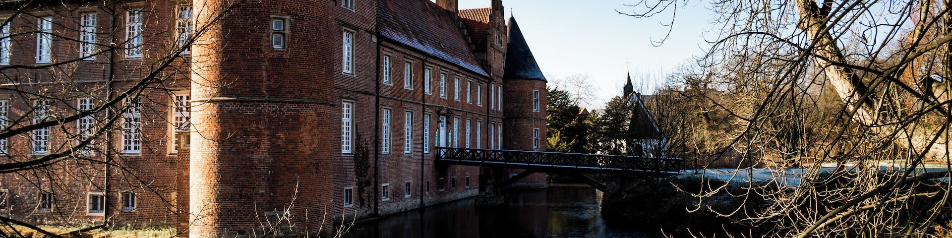 A historic brick building with a tower, a bridge over a river, and leafless trees.