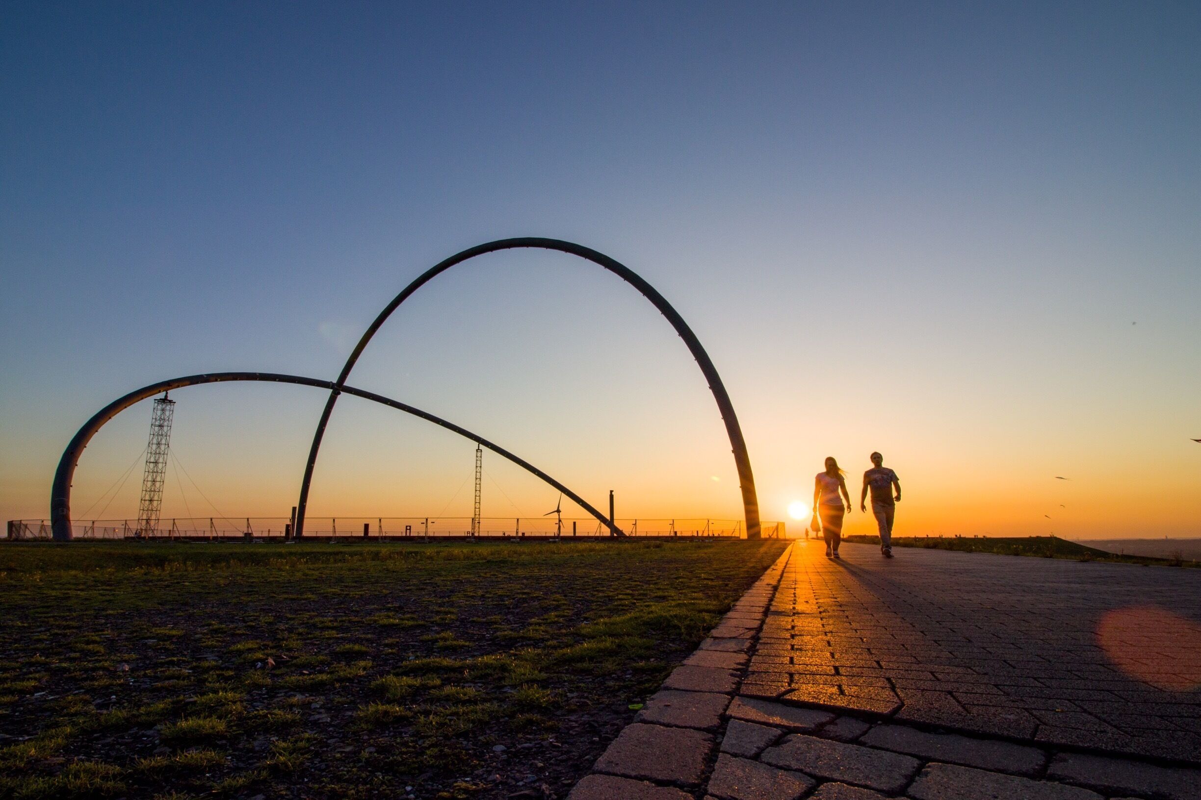 A sunset with two people walking on a paved path, a large arch structure, and wind turbines in the background.