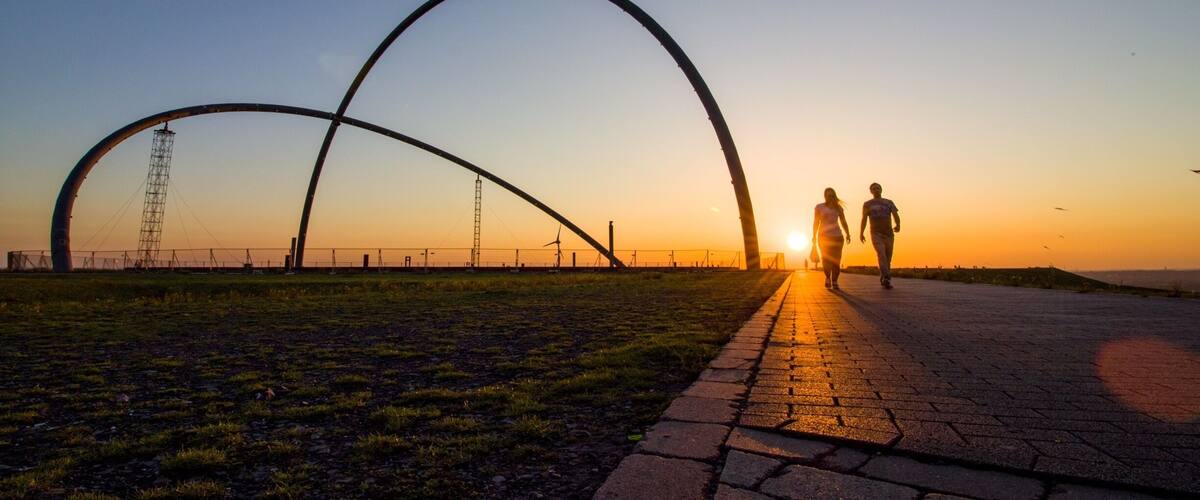 A sunset with two people walking on a paved path, a large arch structure, and wind turbines in the background.