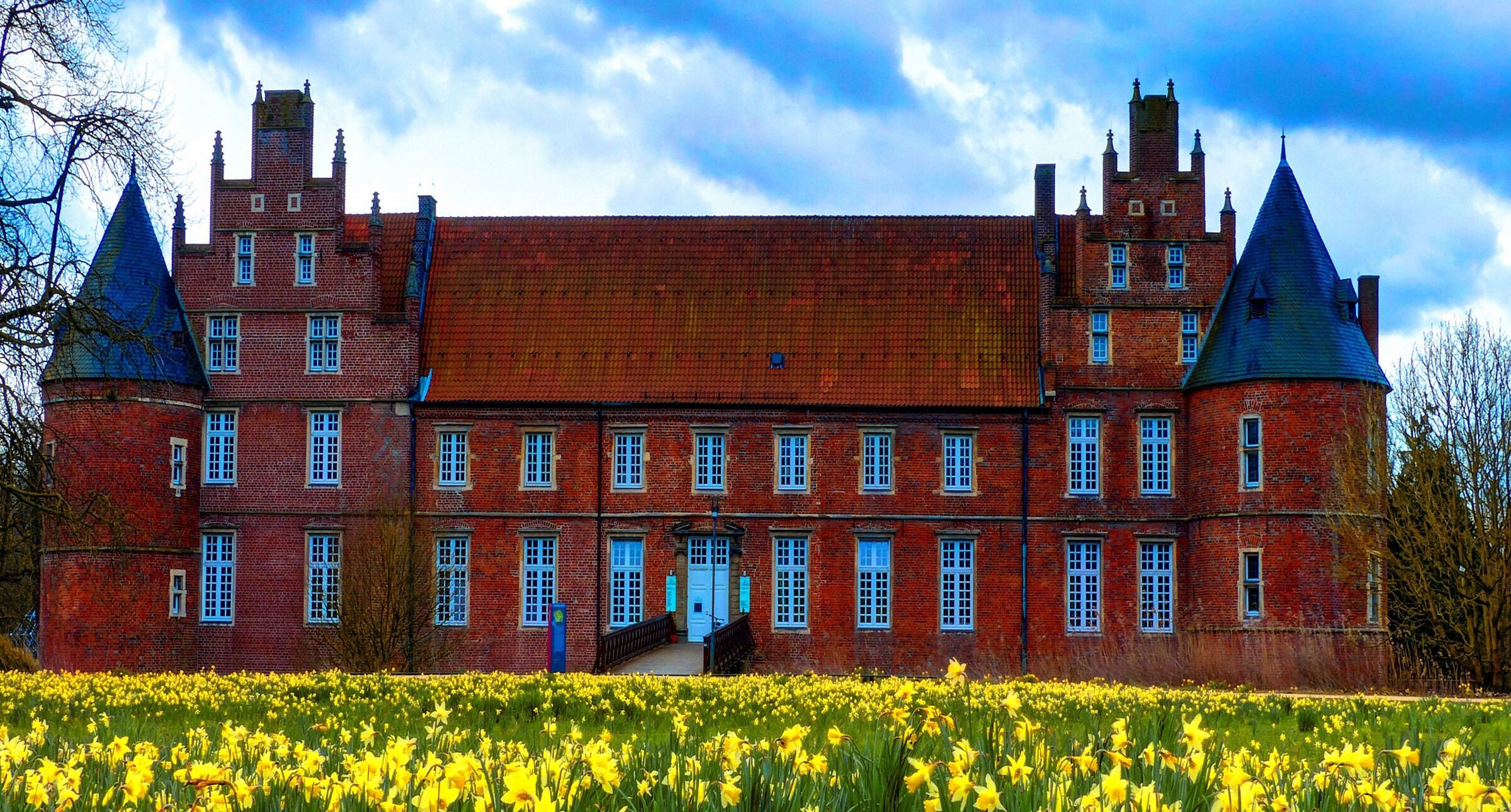 A large red brick building with multiple towers and a field of yellow flowers in front.