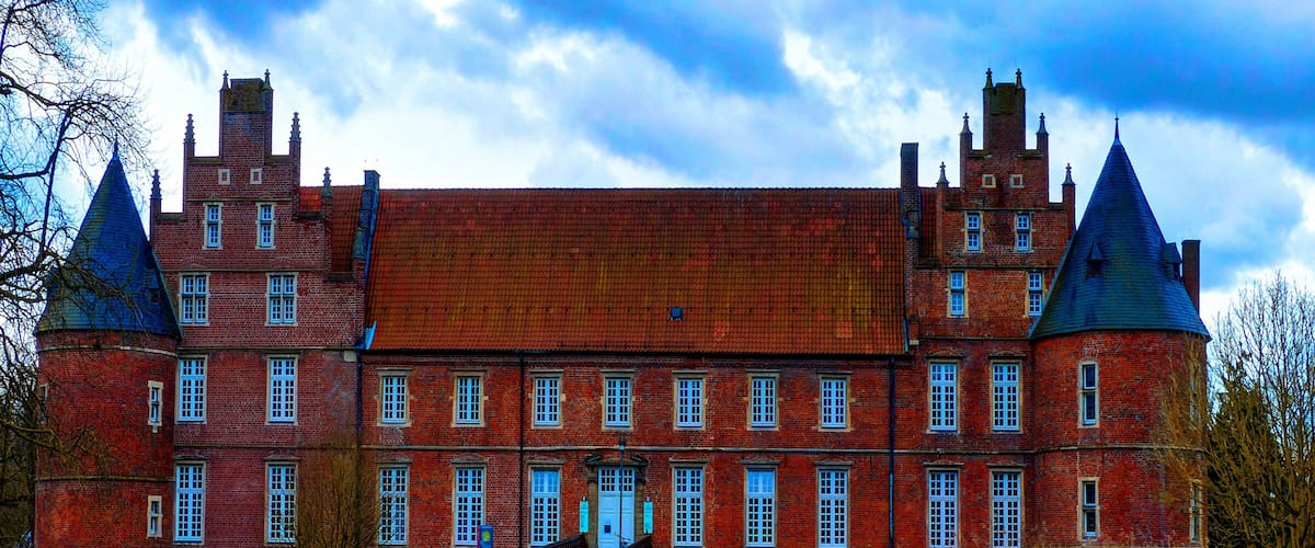 Un grand bâtiment en briques rouges, doté de nombreuses tours, et un champ de fleurs jaunes devant.