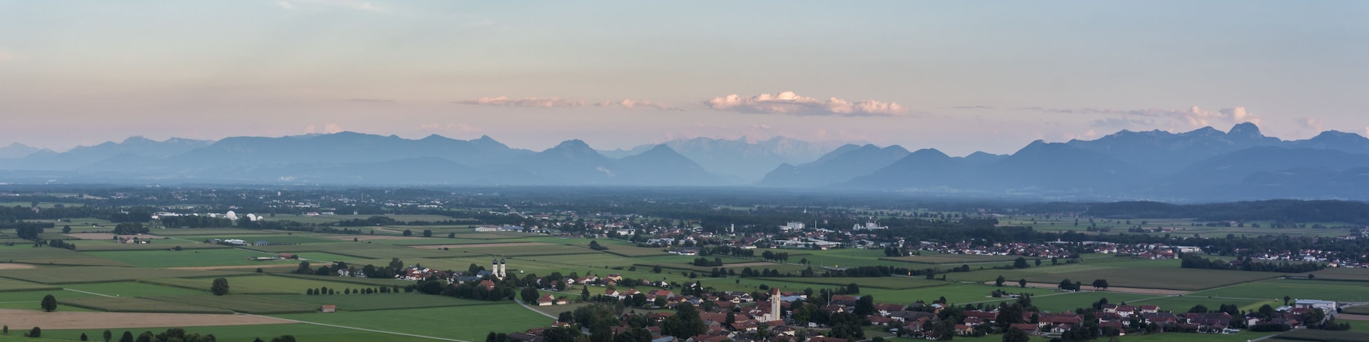 Paisaje rural con colinas onduladas, un pueblo pequeño y montañas a lo lejos.