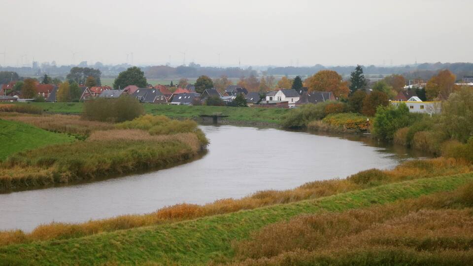 A river meandering through a rural landscape with houses and wind turbines in the distance.
