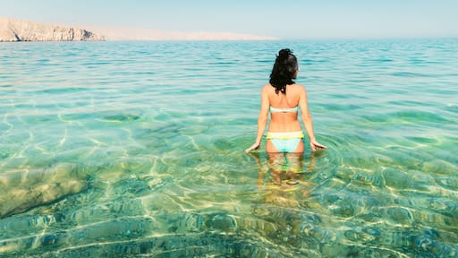 Back view pretty tourist woman stand in clear turquoise water in persian gulf Mirellas island. Oman coastline paradise. Musandam.