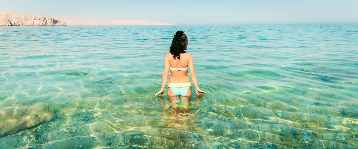 A person standing in clear, shallow water with a distant rocky coastline.