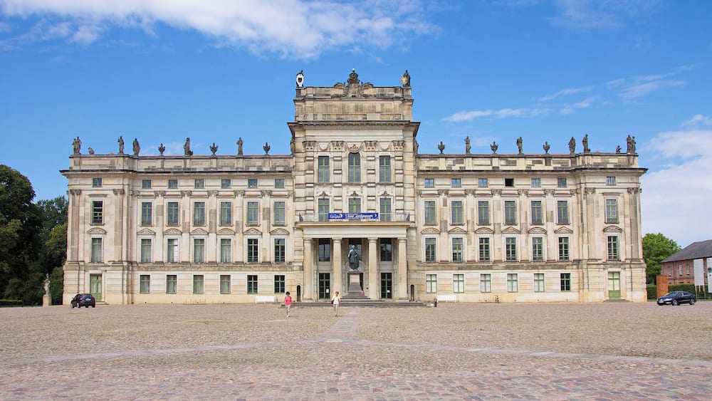 Un bâtiment historique orné de statues sur le toit, doté de grandes fenêtres et d’une cour dallée.