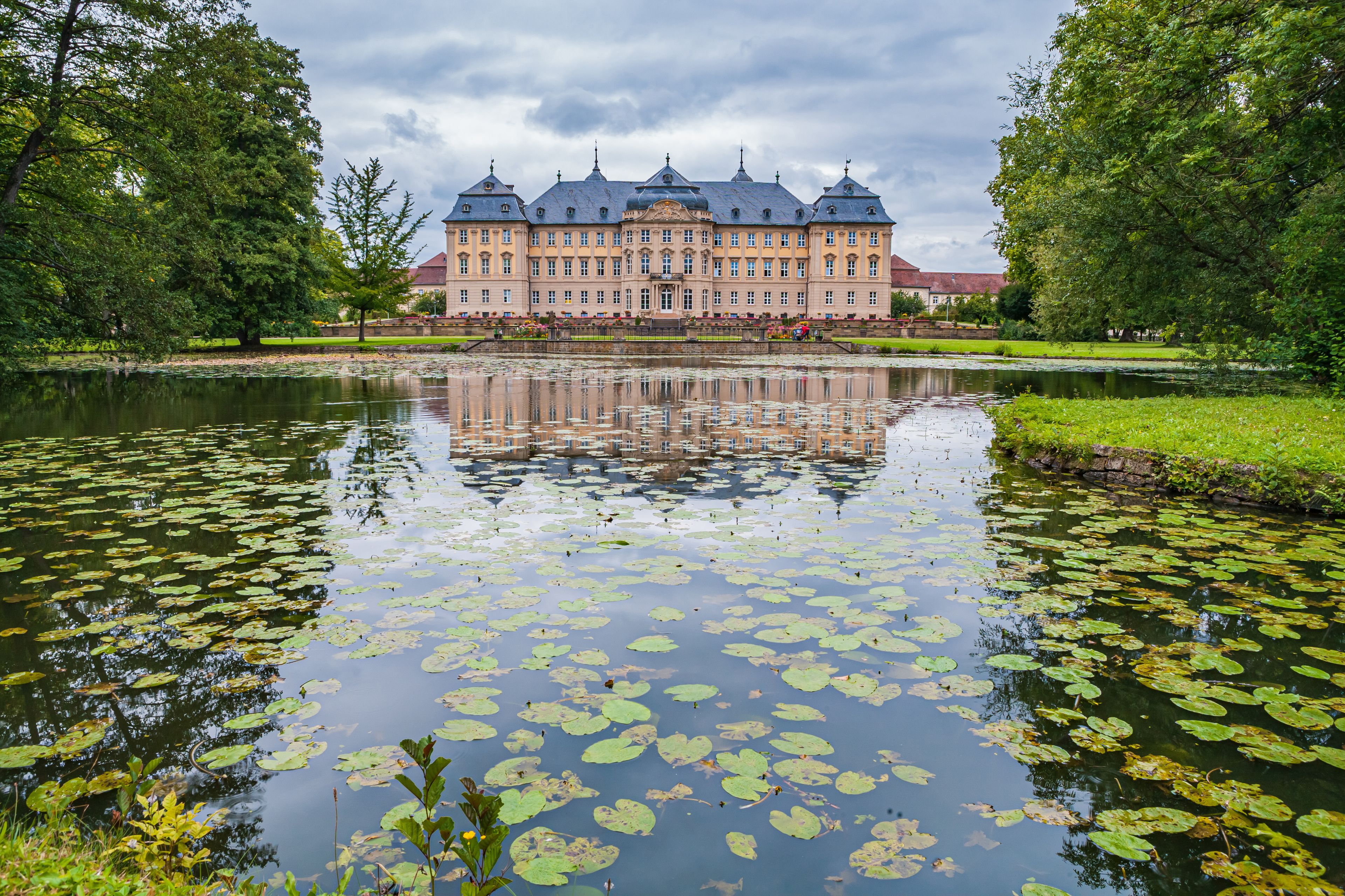 A large, historic building with a symmetrical facade and multiple towers, surrounded by a calm pond with lily pads and lush greenery.
