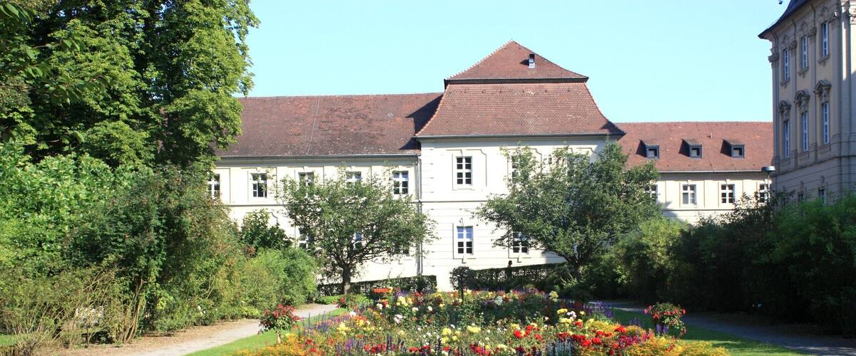 A large white building with a red roof, surrounded by a well-maintained garden with various colorful flowers and trees.