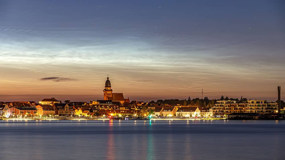 Ein Stadtbild im Abendlicht mit einem markanten Turm und beleuchteten Gebäuden entlang eines Ufers.