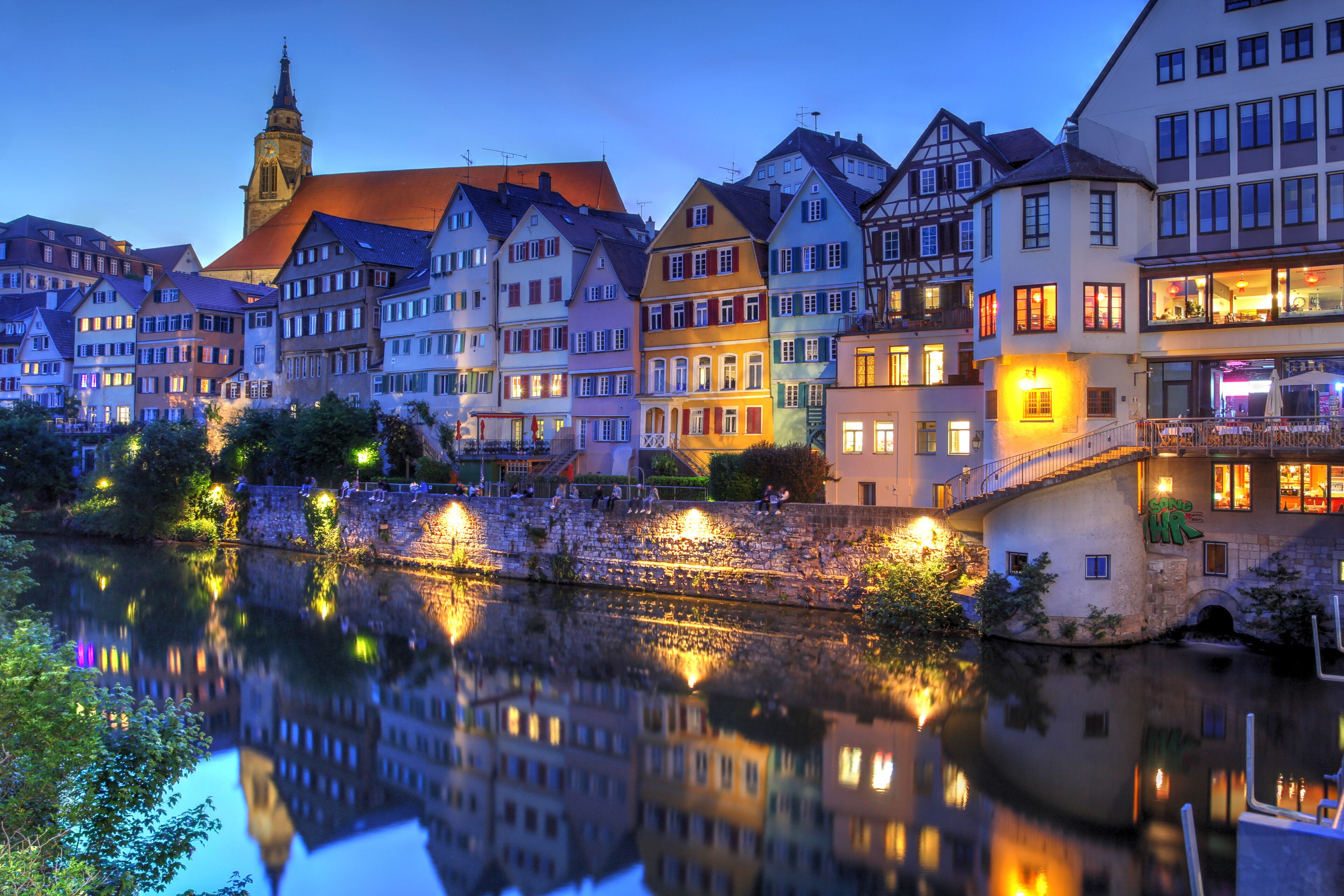 A river reflecting buildings, a church steeple, and a stone bridge.