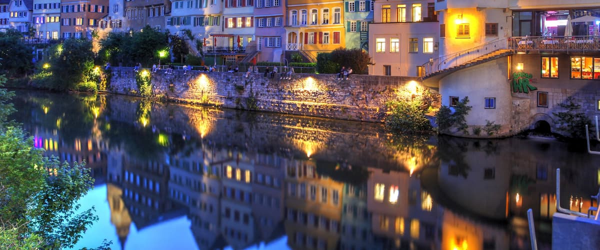 A river reflecting buildings, a church steeple, and a stone bridge.