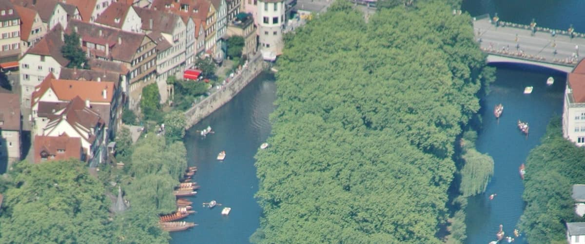 A river flanked by dense green trees and historic buildings with red roofs.