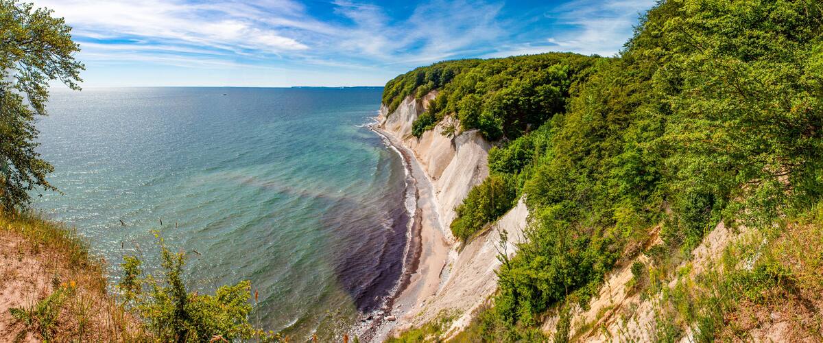 A coastal landscape with a sandy beach, a clear blue sea, and a forested cliff.