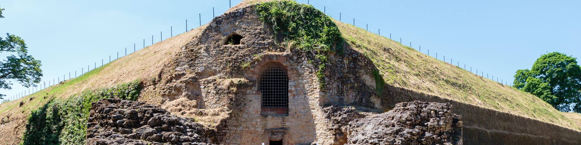 An ancient stone structure with a staircase leading to a central arched entrance, surrounded by a grassy area and a metal fence.