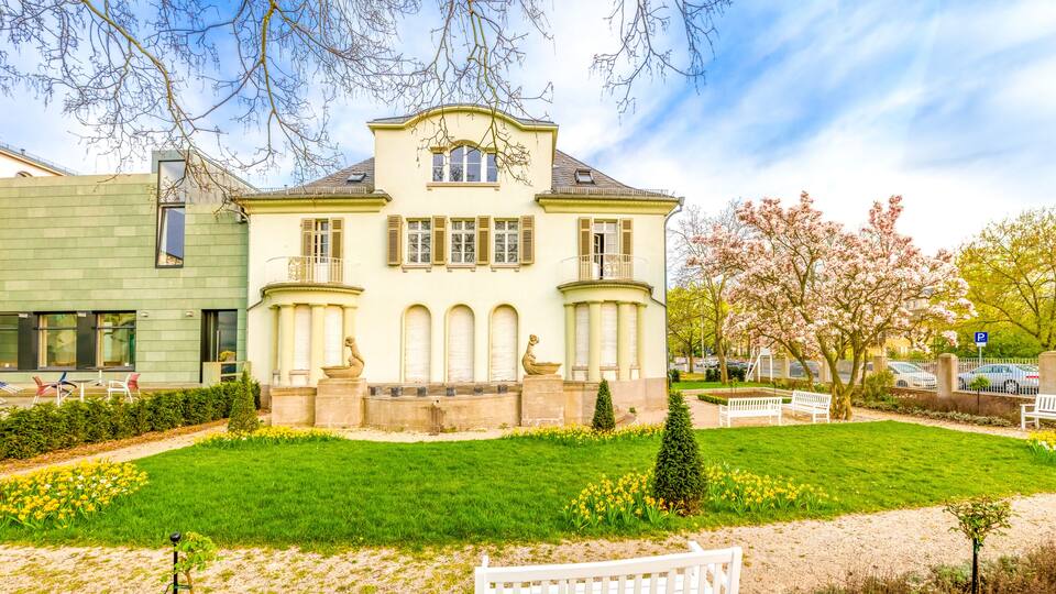 A large, historic building with a green facade, surrounded by a well-maintained garden with flowers and a white bench.
