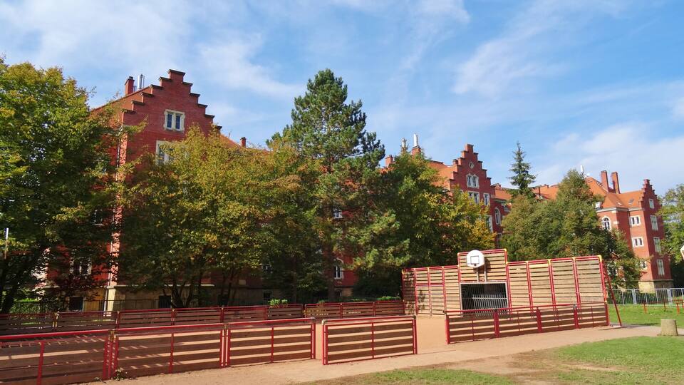 A park with a basketball court, red fences, and a building with red brick architecture.