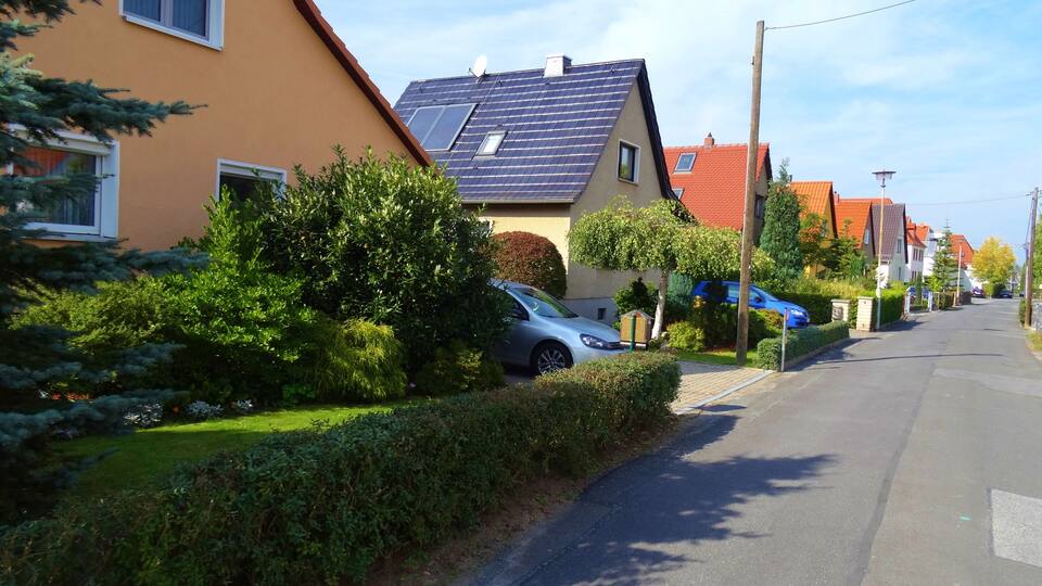 A residential street with houses, a parked car, and lush greenery.