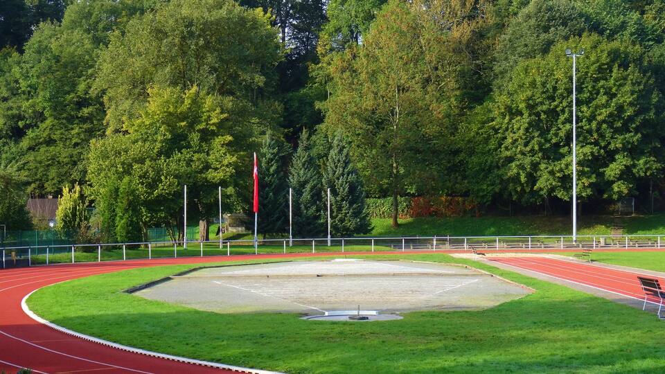 A running track with a red surface, surrounded by green grass and trees.