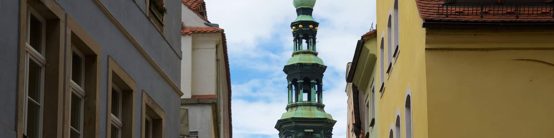 A narrow street with buildings on either side, a green clock tower, and a clear sky.