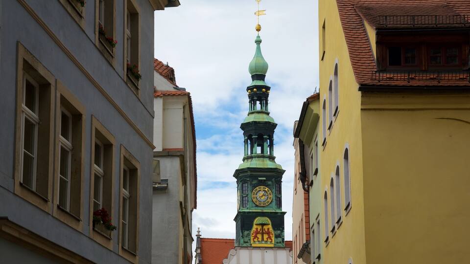 A narrow street with buildings on either side, a green clock tower, and a clear sky.