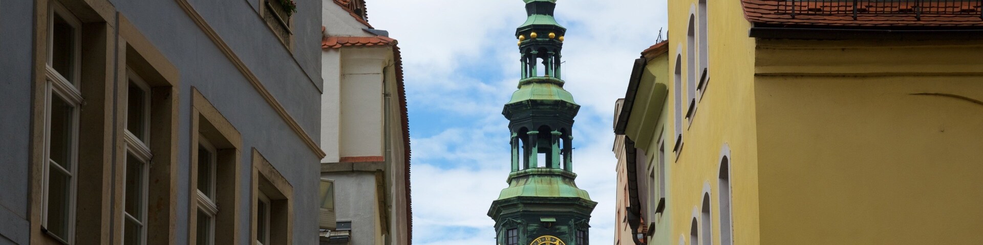 Una calle estrecha flanqueada por edificios, una torre del reloj verde y un cielo despejado.