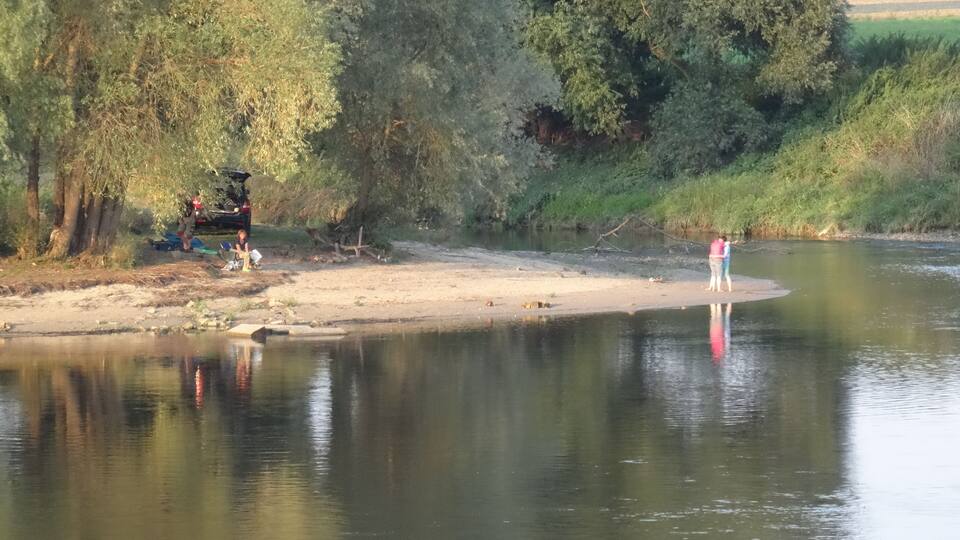 A riverbank with people near the water, trees, and a distant field.