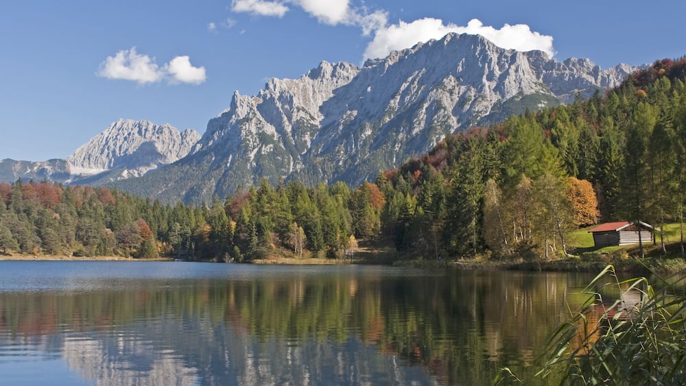 A calm lake with reflections of trees and mountains, surrounded by autumn foliage and a small cabin.